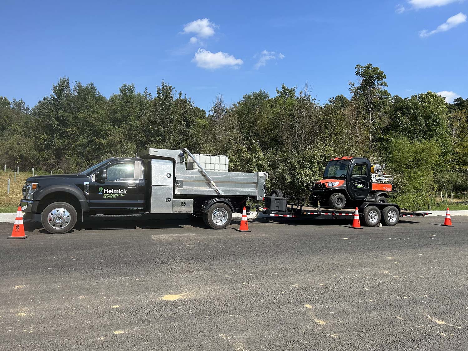 A black truck with a silver utility bed and an orange utility vehicle on a trailer, surrounded by traffic cones, against a green backdrop.