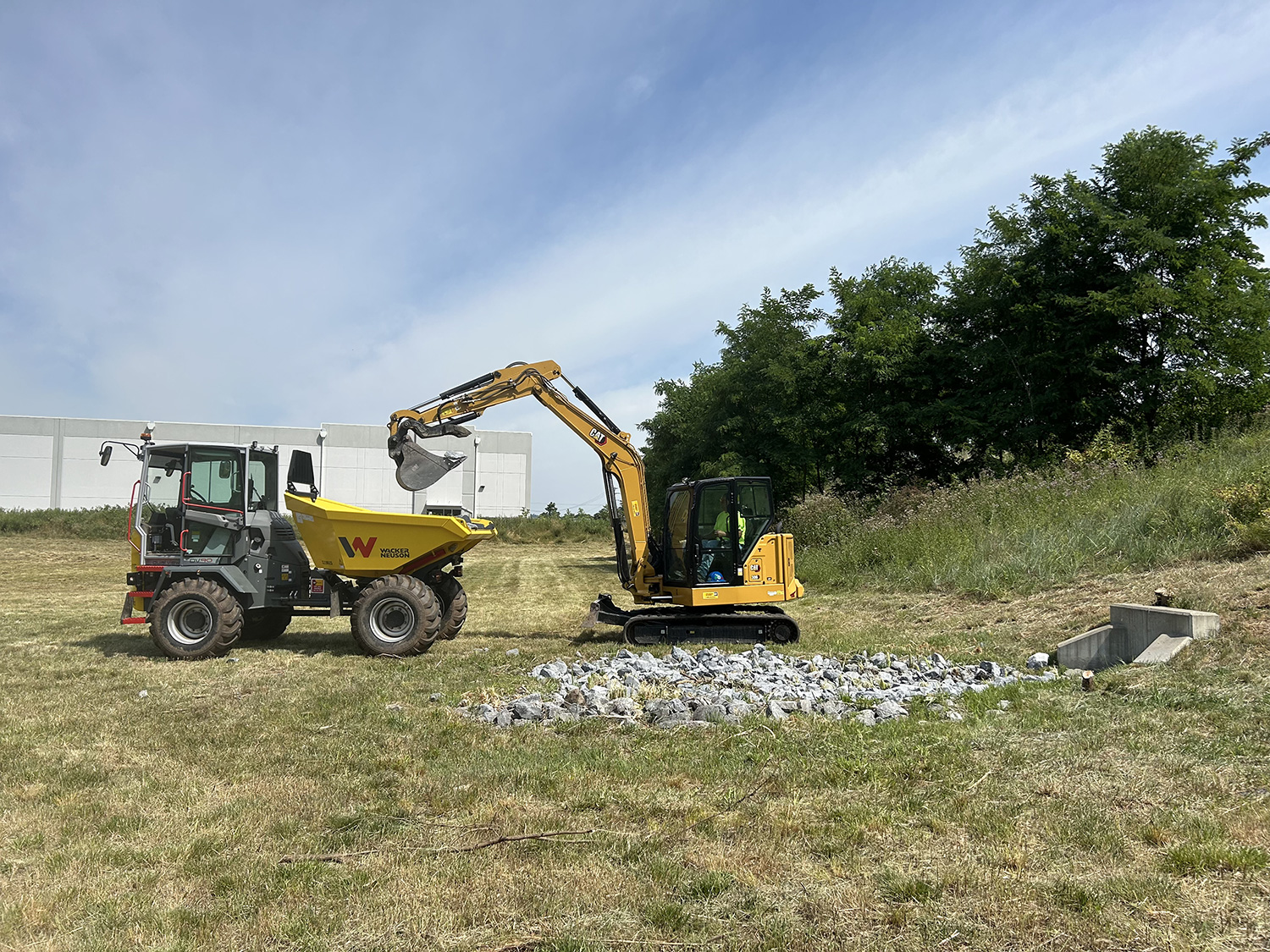 A yellow excavator lifts rocks while a wheeled dumper truck waits nearby on a grassy construction site under a clear sky.