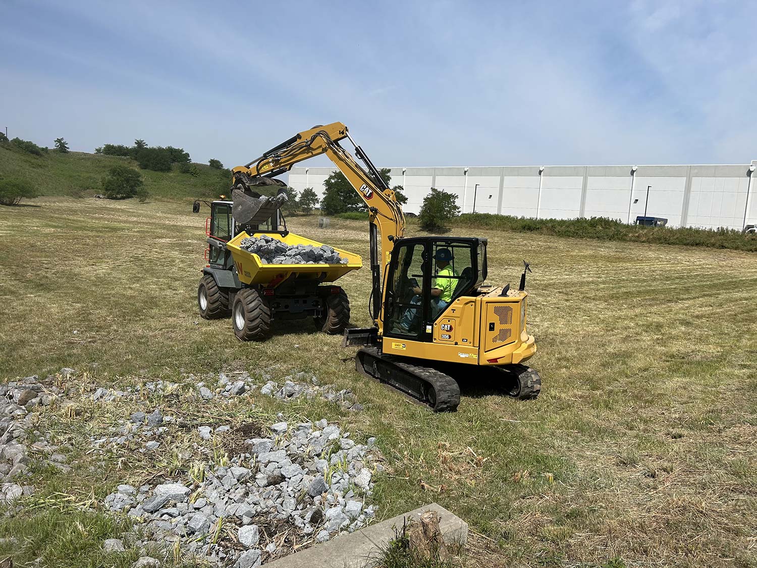 A yellow excavator loads rocks into a yellow dumper truck on a grassy field, with a warehouse visible in the background.