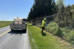 A worker in safety gear sprays vegetation near a chain-link fence, while a liquid control truck is parked on a sunny roadside.