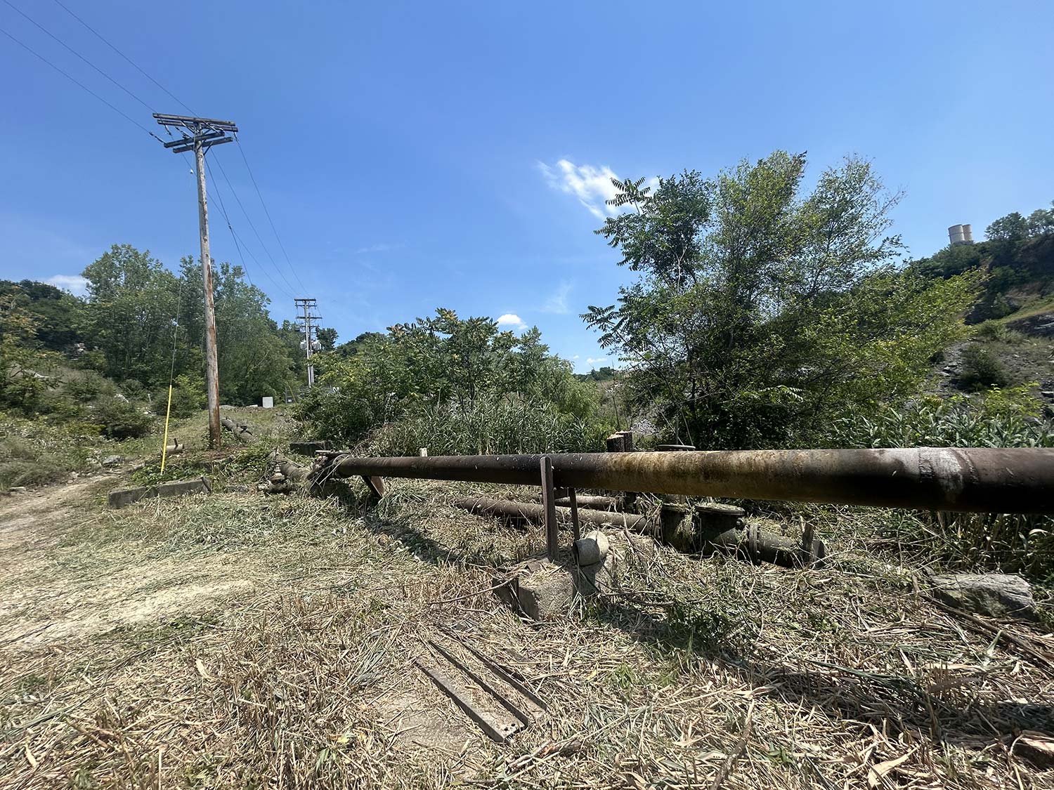 A sunlit landscape features a rusty pipeline alongside electric poles, surrounded by greenery and blue skies.