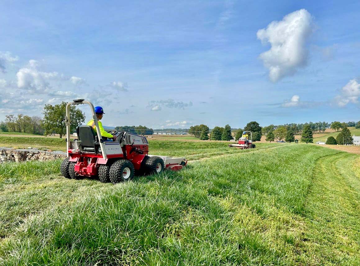 A worker mows grass on a riding lawnmower in a lush green field under a blue sky with scattered clouds, while another mower is in the distance.