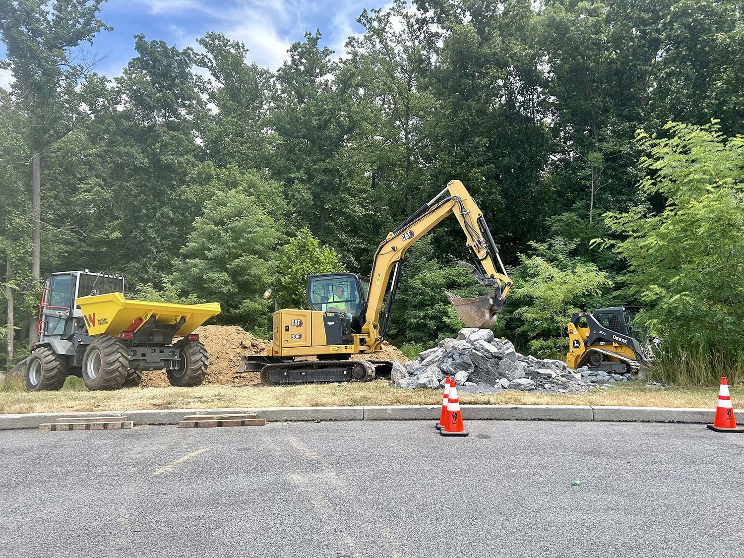Construction site with three heavy machines: a dump truck, an excavator lifting rocks, and a skidded steer loader near trees.