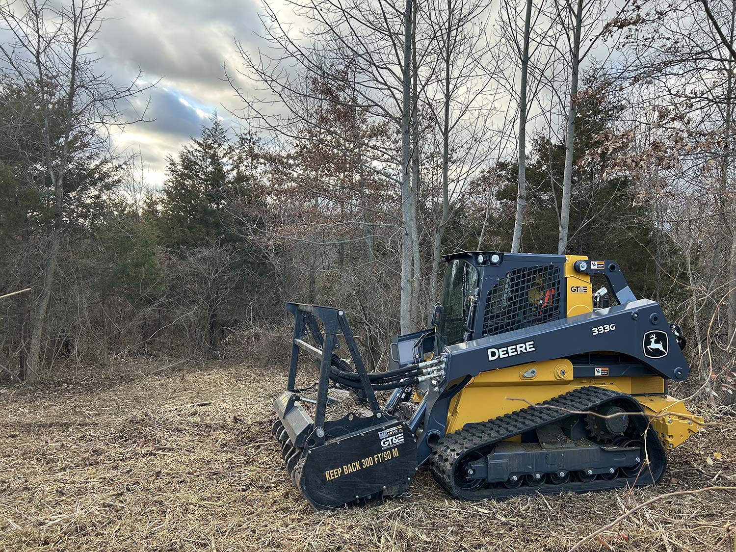 A John Deere 333G compact track loader stands in a wooded area, with bare trees and cloudy skies in the background.