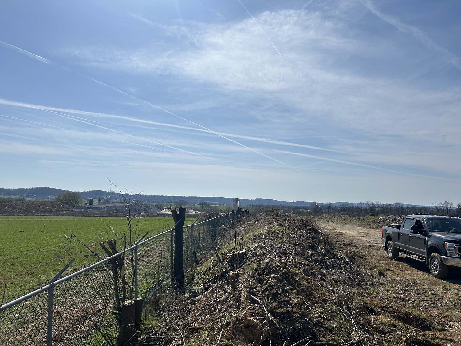 A clear blue sky with contrails stretches over a grassy field, a wooded area, and a parked truck beside a chain-link fence.