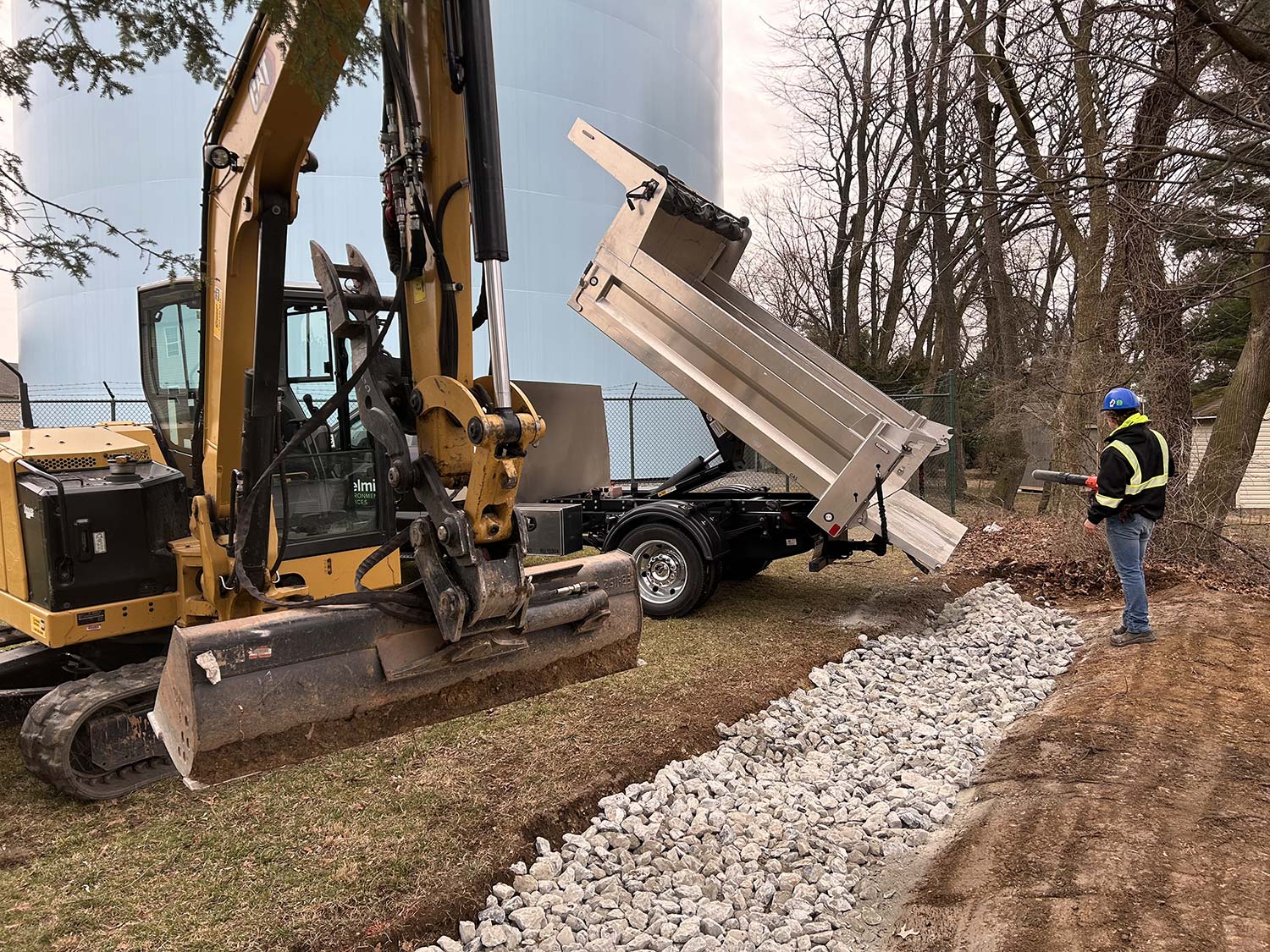 A dump truck unloads gravel while a worker observes, with heavy machinery and trees in the background, near a storage facility.
