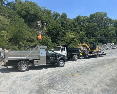 Two work trucks and a mini excavator are parked on a gravel site, surrounded by trees and rocky terrain under a clear blue sky.