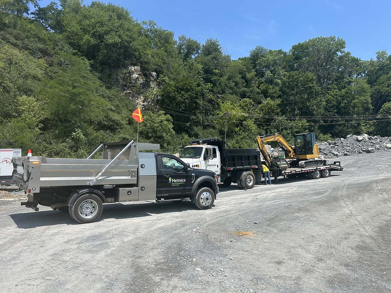 Two work trucks and a mini excavator are parked on a gravel site, surrounded by trees and rocky terrain under a clear blue sky.