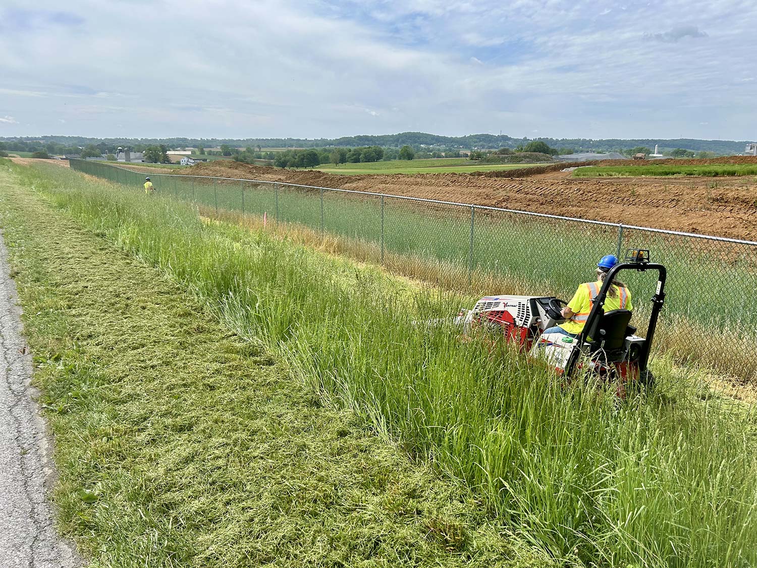 A worker mows tall grass along a chain-link fence, with green fields and a blue sky in the background.