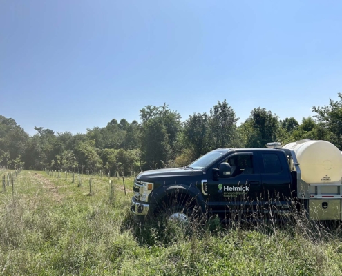 A black truck with a Helminck logo parked in a grassy field lined with young trees under a clear blue sky.