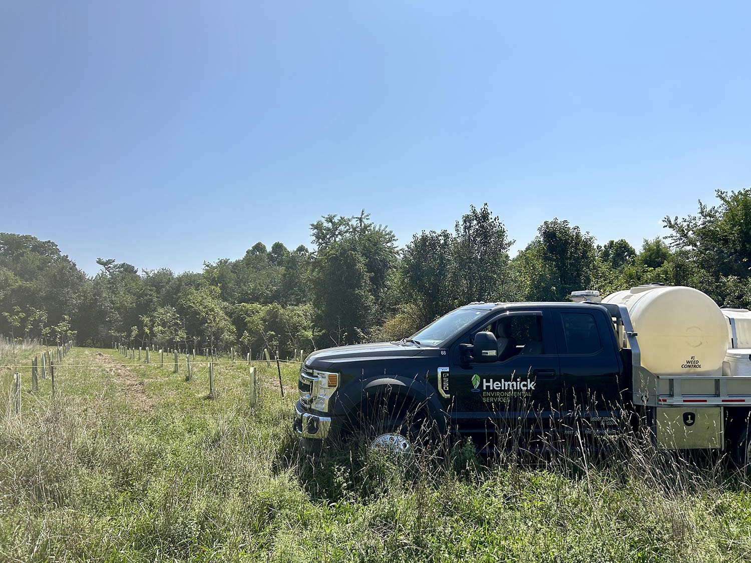 A black truck with a Helminck logo parked in a grassy field lined with young trees under a clear blue sky.