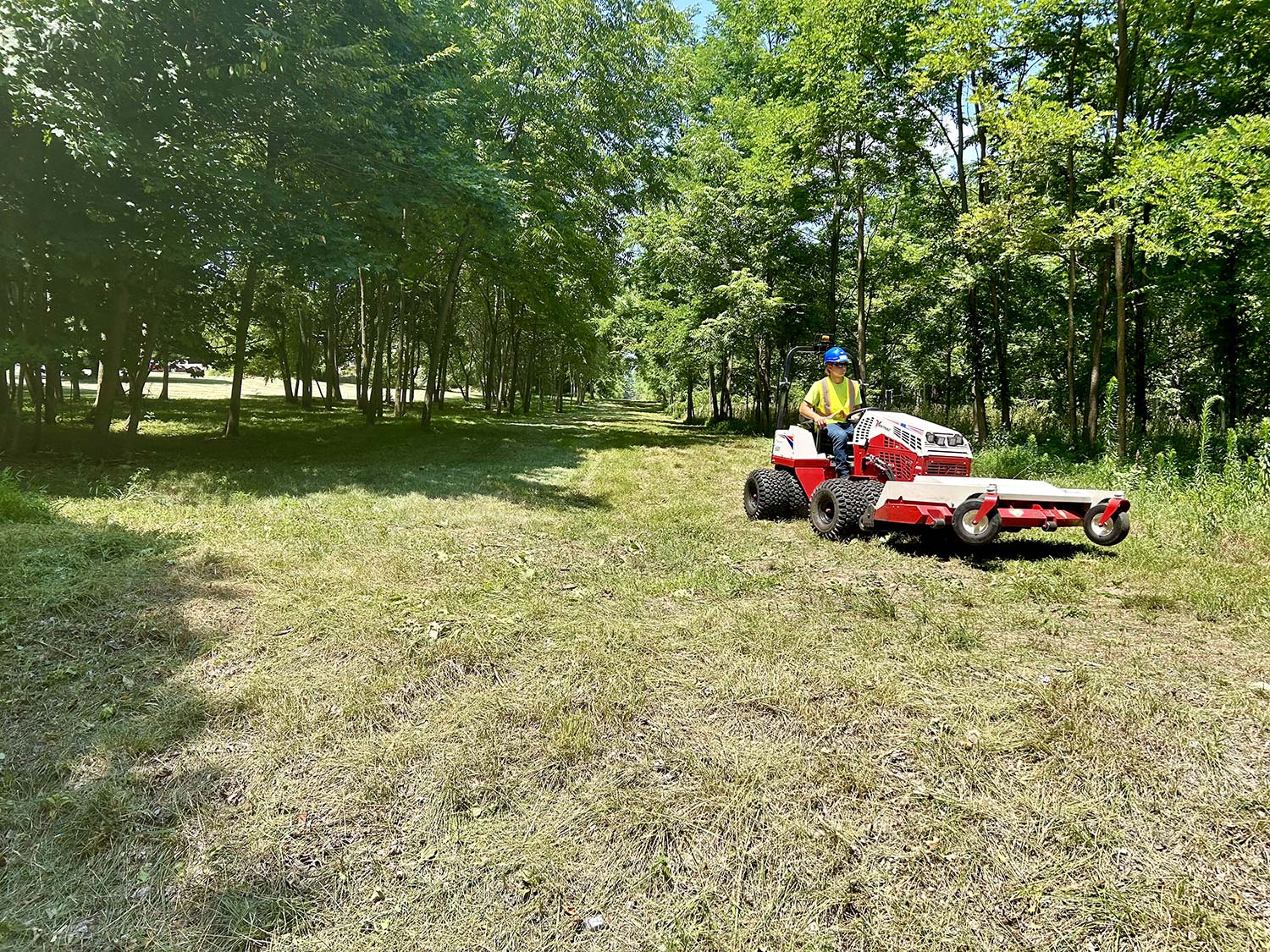 A worker in a yellow safety vest operates a riding lawn mower, cutting grass along a wooded pathway on a sunny day.