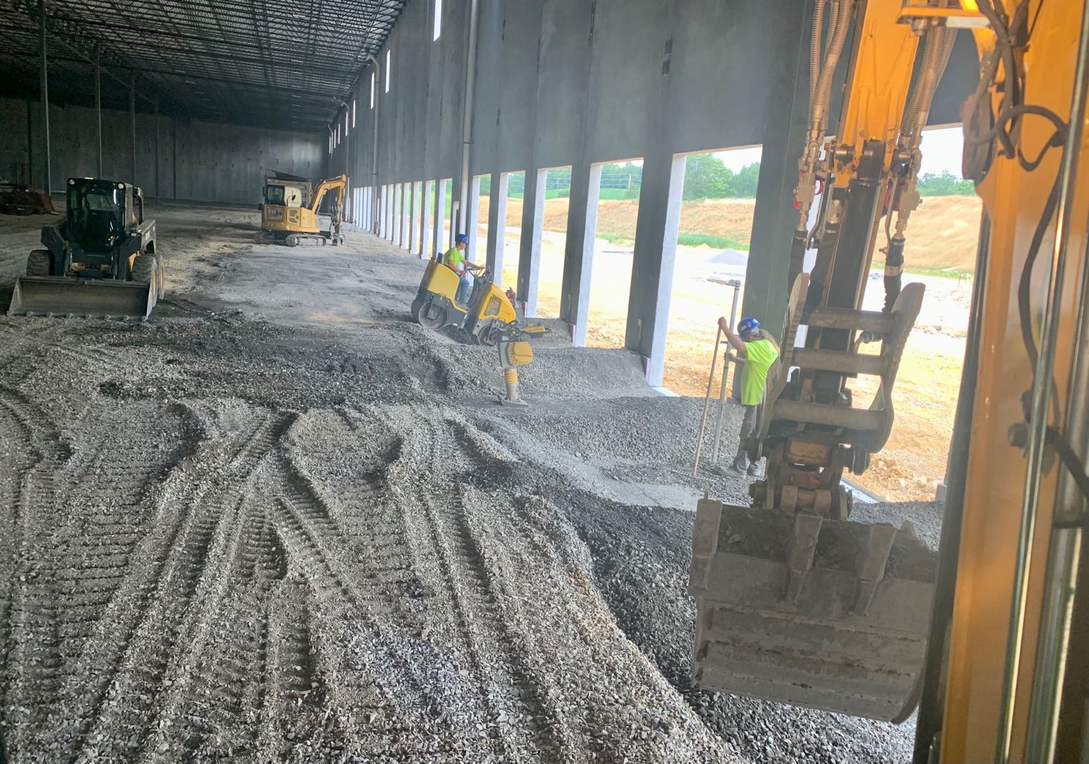 Workers operate heavy machinery inside a large warehouse, spreading gravel and compacting the floor under natural light from windows.
