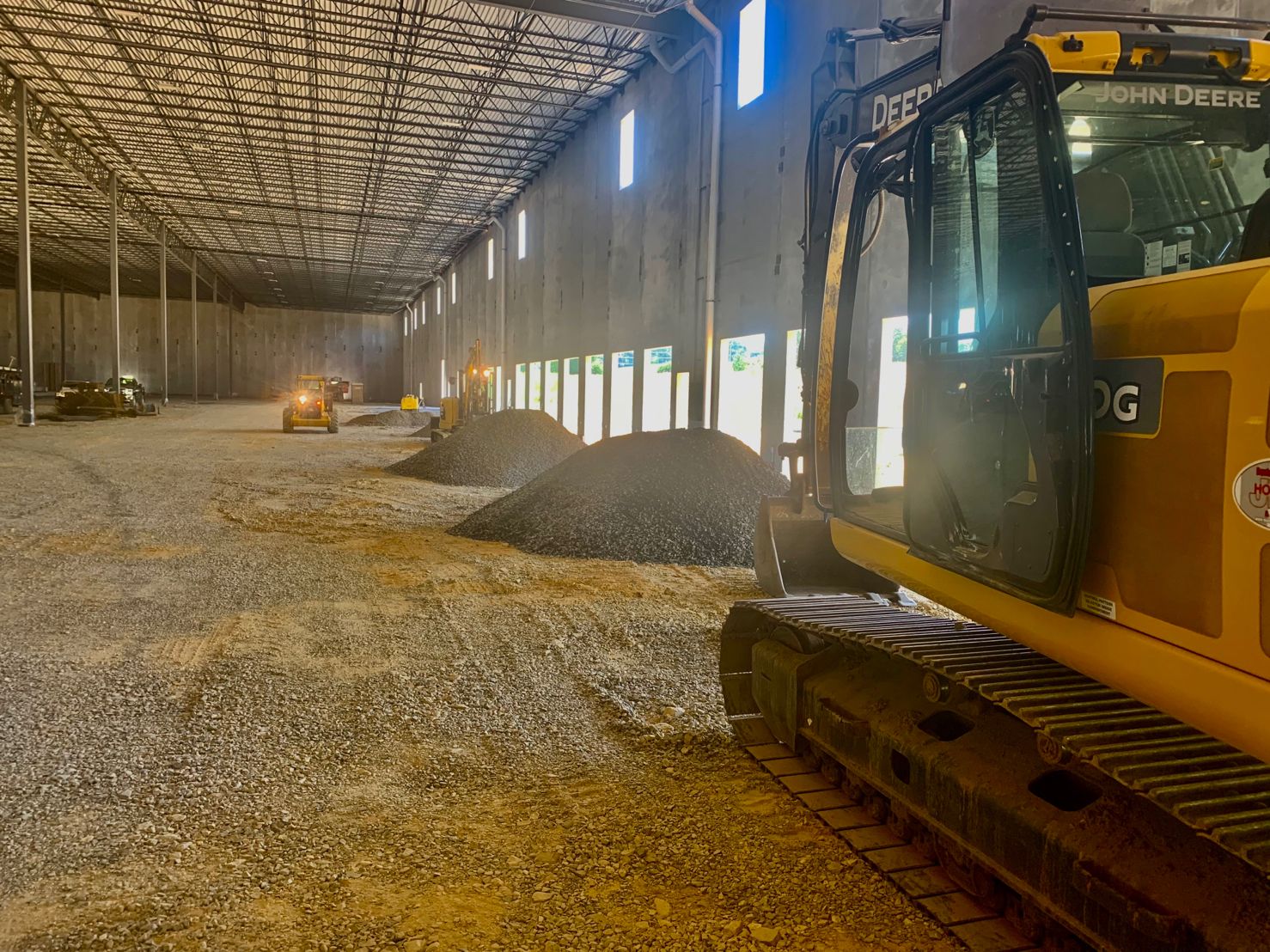 Inside a large warehouse, heavy machinery is parked near piles of gravel, with bright windows lining the wall.