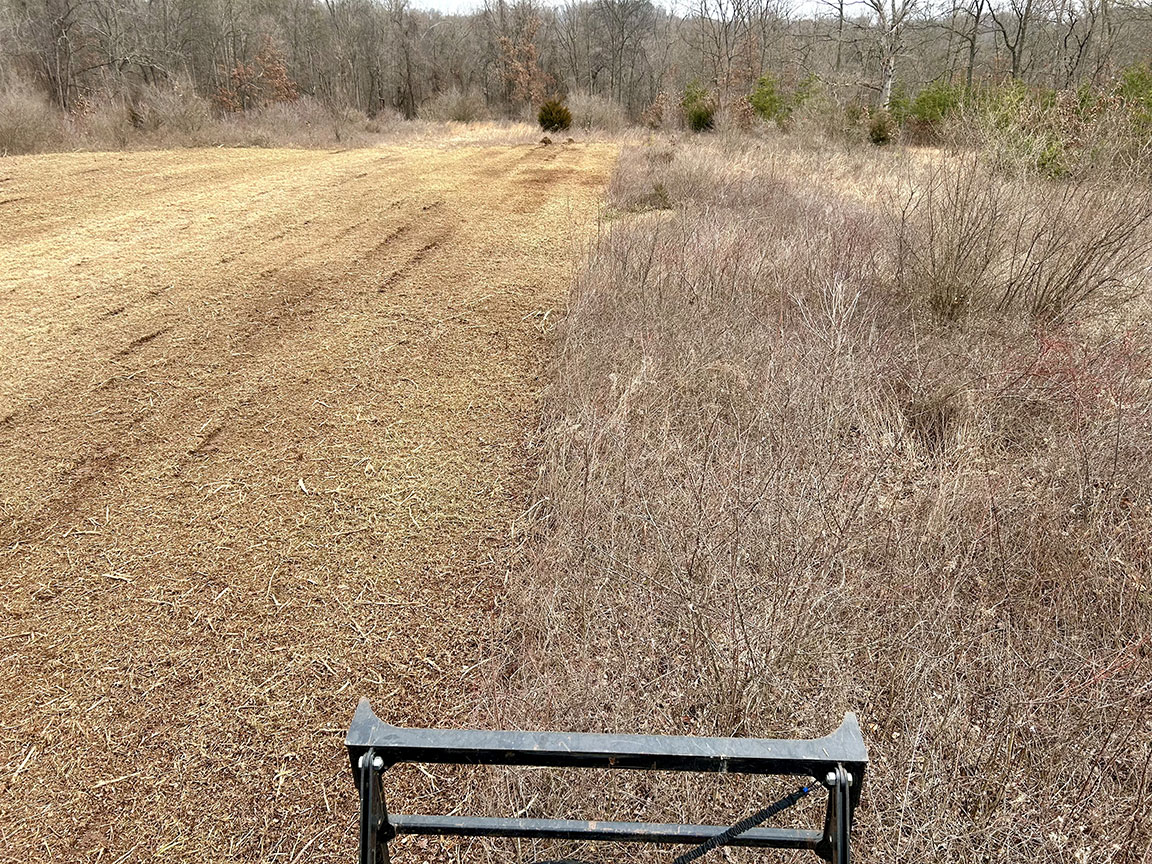 A cleared, brown field stretches before dense, dry underbrush and trees, revealing remnants of greenery and a machine's edge in the foreground.