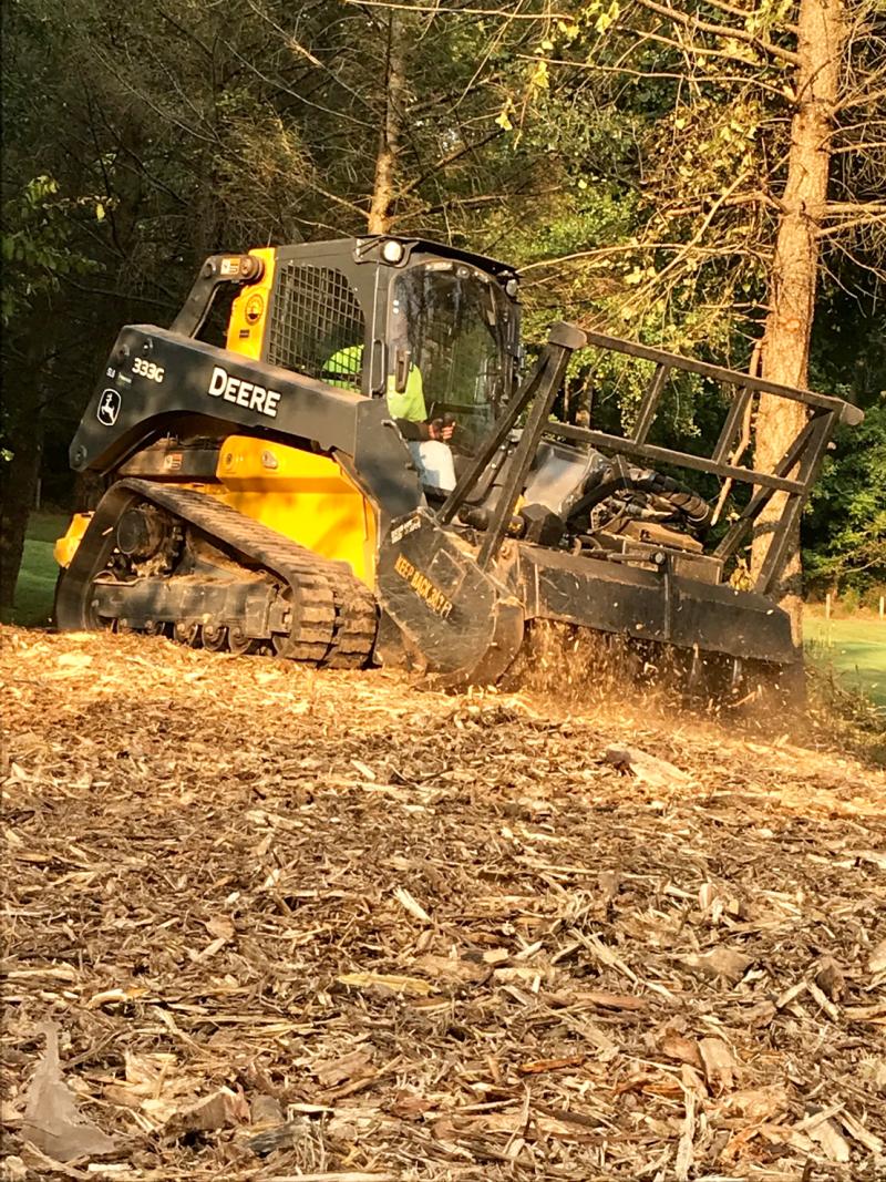A John Deere 333G skid steer works on a wooded area, churning up wood chips and debris under a sunny sky.