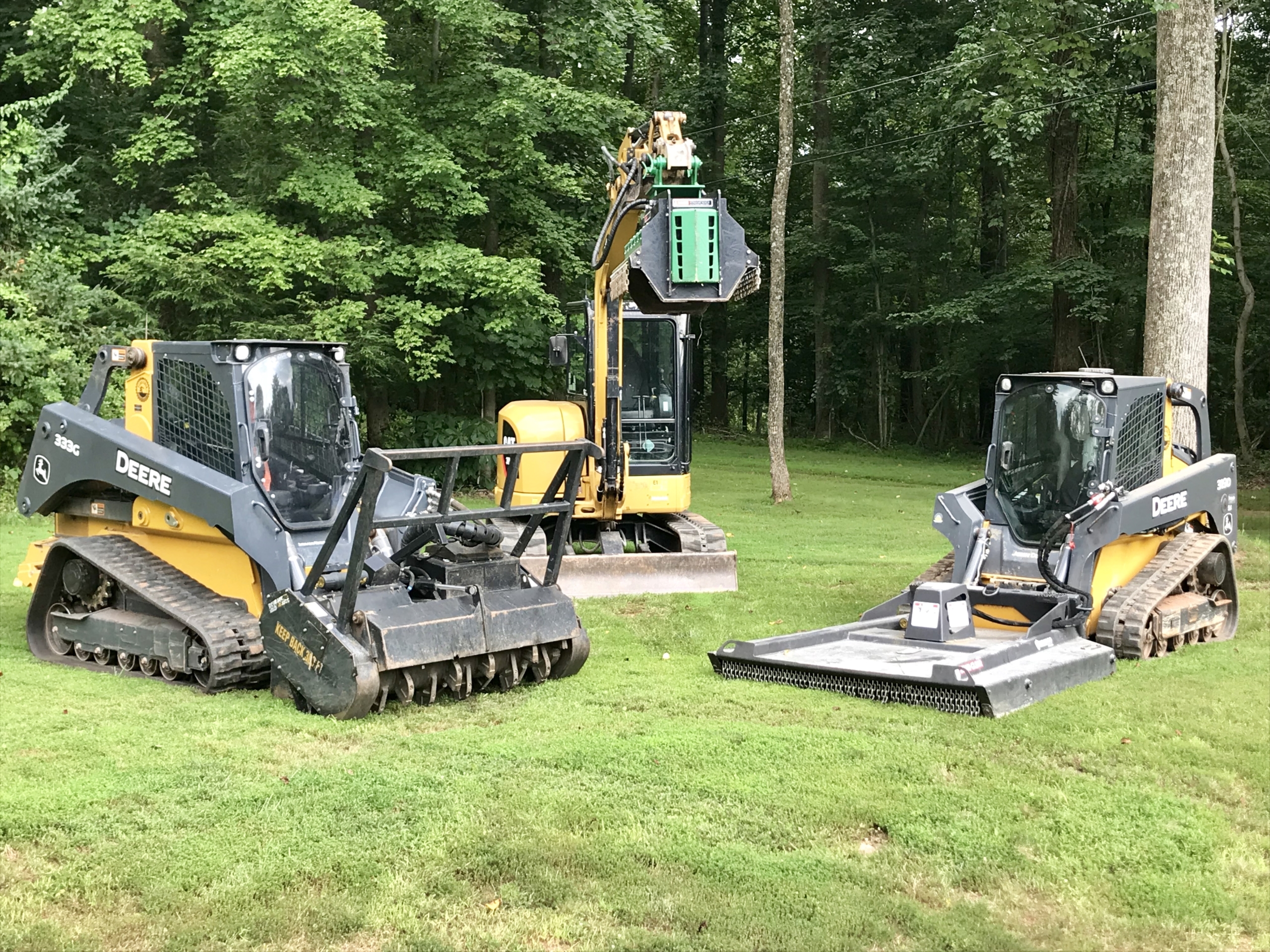 Two yellow and black John Deere skid steer loaders and a larger excavator are parked on green grass surrounded by trees.